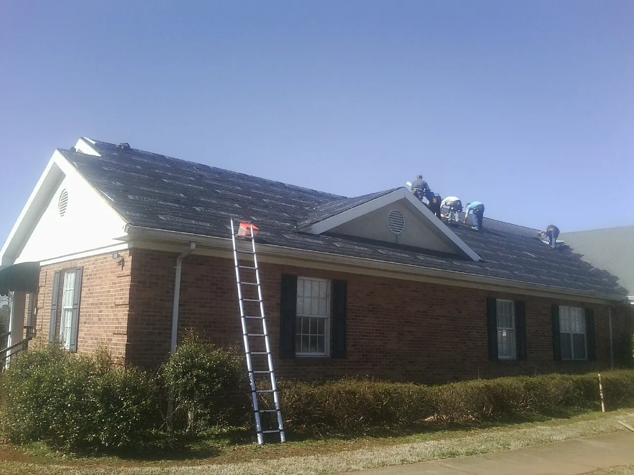 Crew tearing shingles off a brick ranch roof with a ladder set — Metal Roof Repair work by our crew in Bassfield