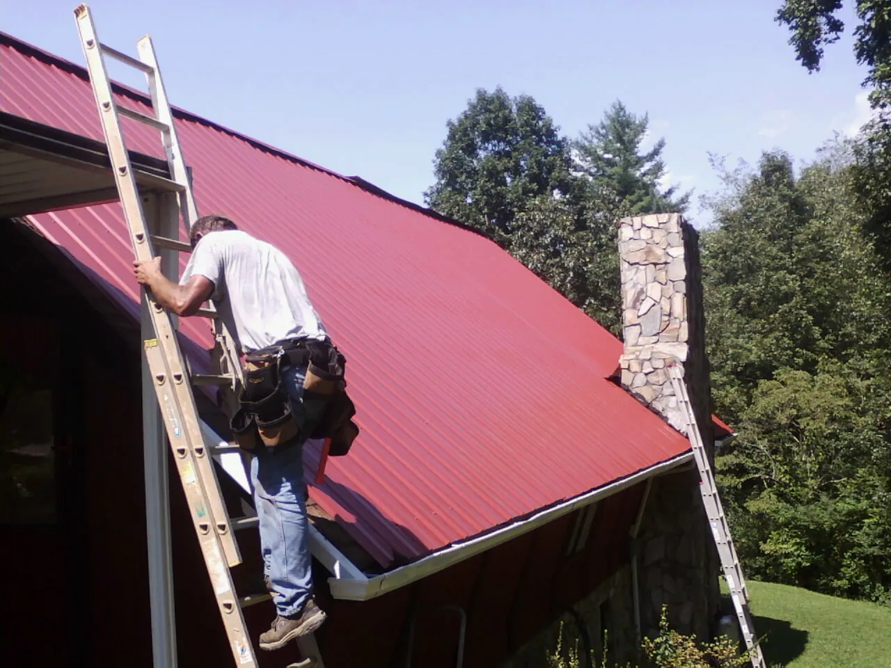 Roofer on a ladder beside a red metal roof and stone chimney — Soffit & Fascia Repair by Roofing Brothers in Bassfield